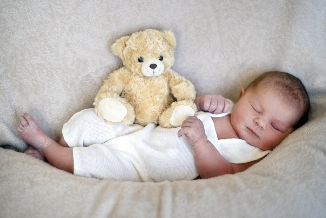 Schlafendes Neugeborenes in weißem Strampler mit Teddybär auf weicher Decke.