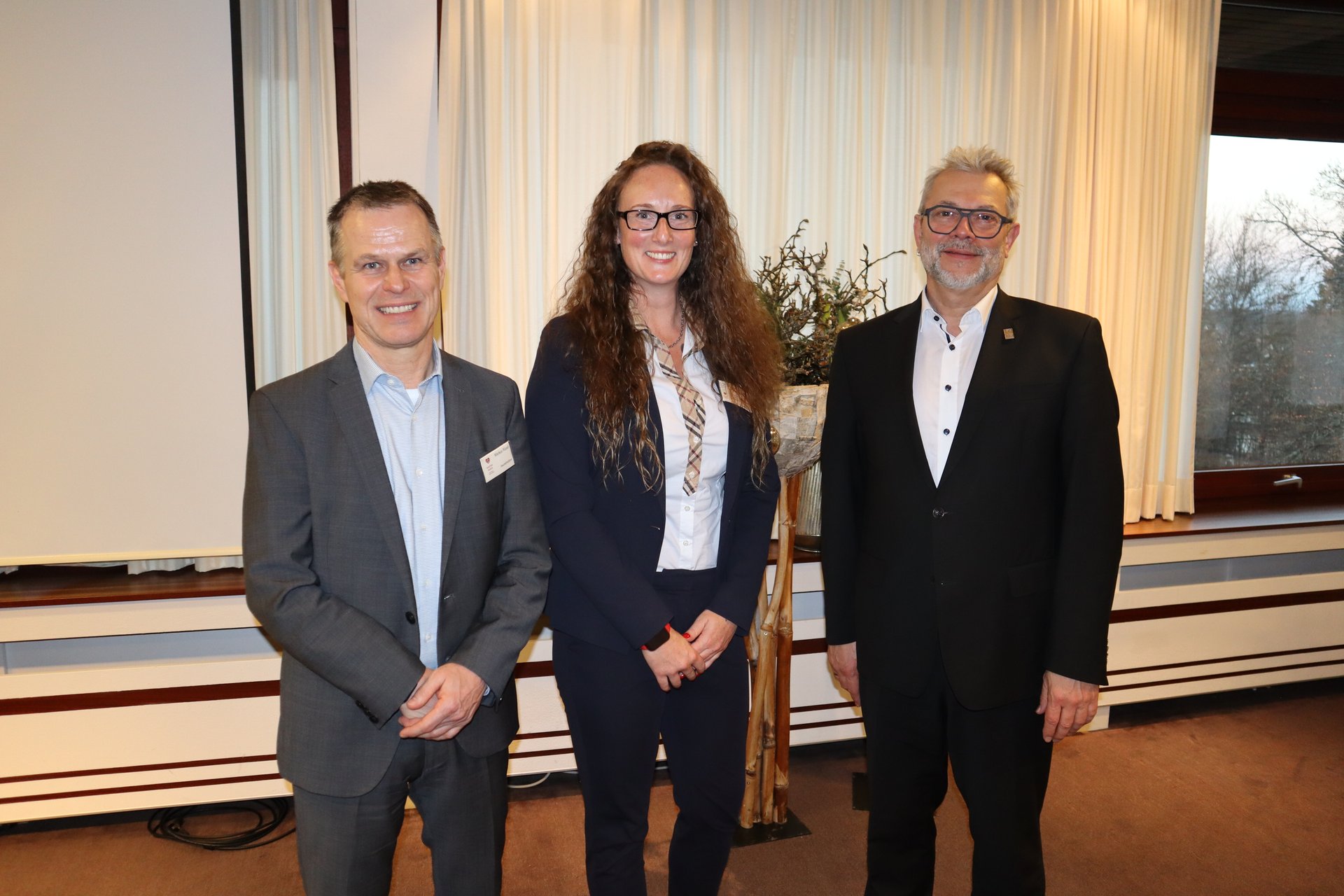 Drei lächelnde Personen in Anzügen posieren vor einem dekorierten Podium.