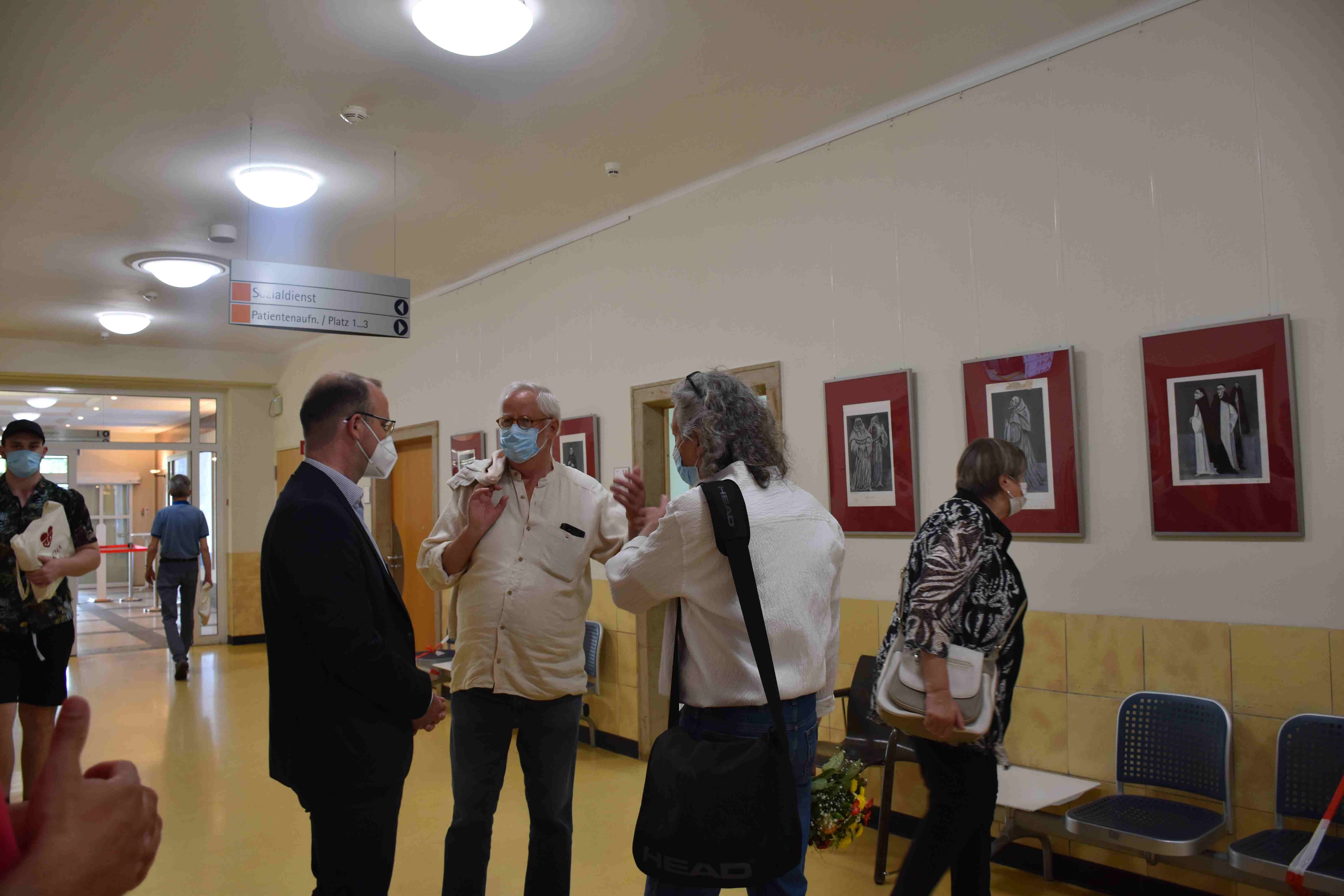 Menschen mit Masken unterhalten sich im Krankenhausflur vor Kunstwerken.