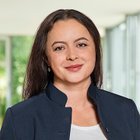 Porträt einer lächelnden Frau in professioneller Kleidung vor Bürohintergrund.