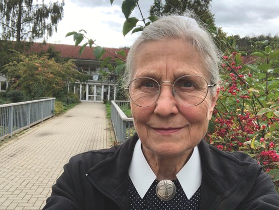 Ältere Dame mit Brille lächelt, umgeben von Natur, vor einem Gebäude.