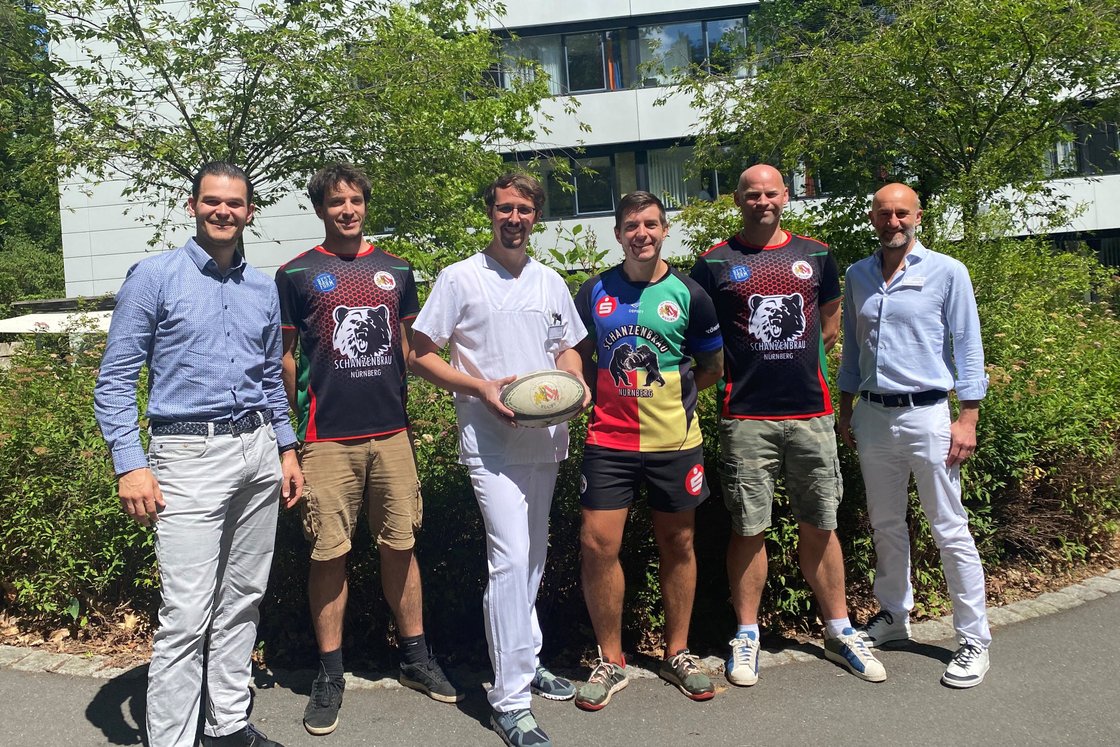 Gruppe von Männern hält Rugbyball vor einem Gebäude.