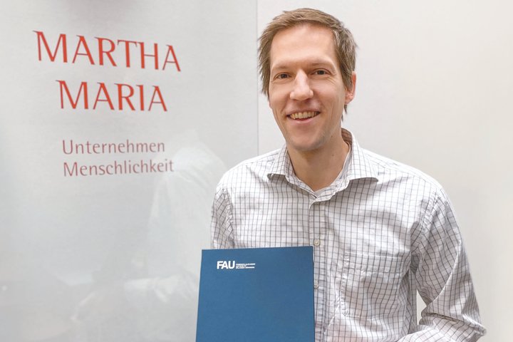 Mann mit FAU-Ordner vor Martha Maria Unternehmensschild lächelnd porträtiert.