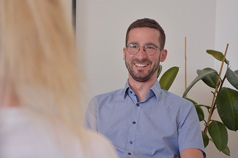 Lächelnder Mann im Gespräch mit einer anderen Person, Pflanze im Hintergrund.