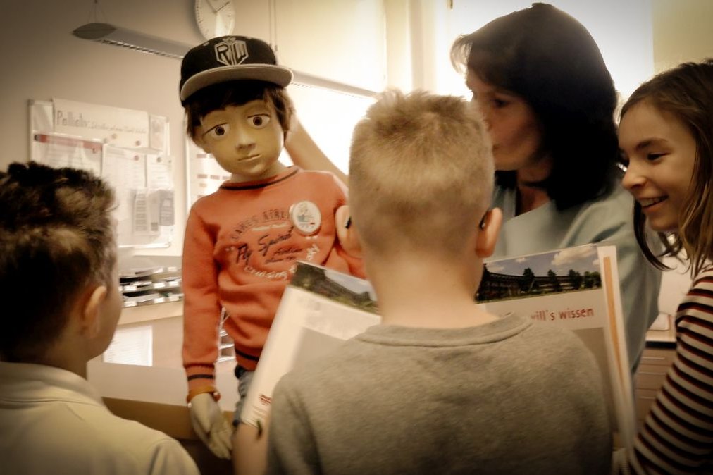 Kinder betrachten Puppe Jonas während eines Krankenhausprojekts, nahe einer Erwachsenen.