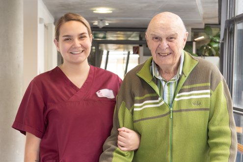 Pflegekraft hilft älterem Mann im Pflegeheim, beide lächeln freundlich.