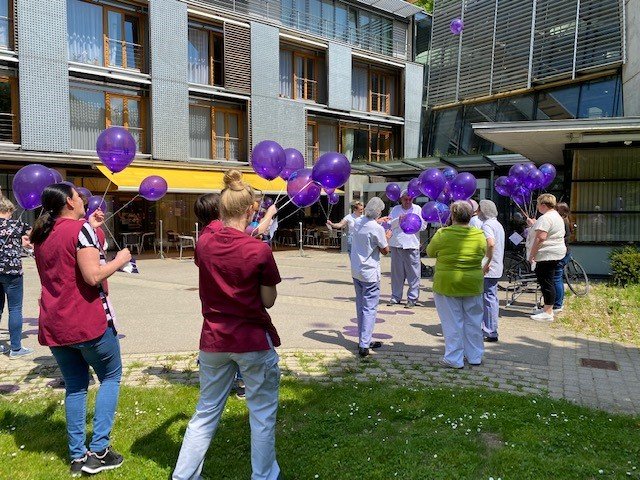 Pflegepersonal lässt lila Luftballons vor einem modernen Gebäude steigen.