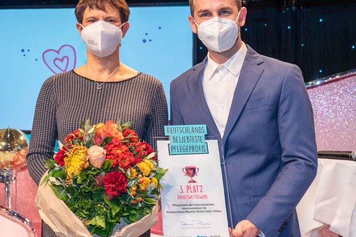 Zwei Personen mit Blumenstrauß und Auszeichnung für Pflegewettbewerb auf Bühne.