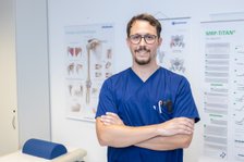 Arzt in blauer Uniform steht lächelnd vor medizinischen Postern in einem Behandlungsraum.
