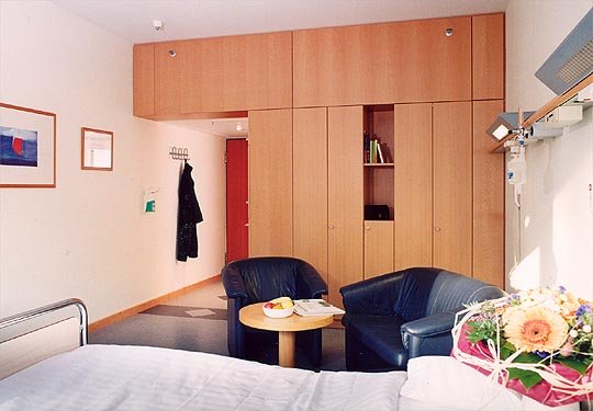 Gemütliches Patientenzimmer mit Bett, Tisch, Sesseln und Blumenarrangement.