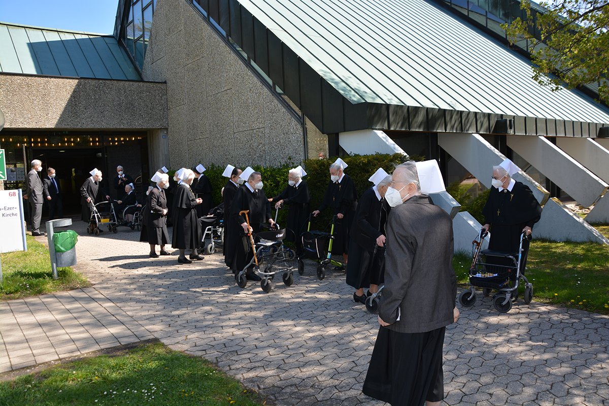 Mehrere Nonnen mit Rollatoren vor einem modernen Kirchengebäude versammelt.