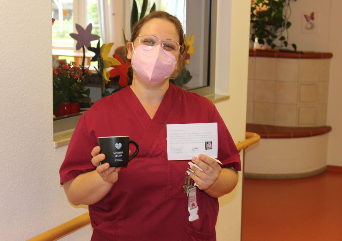 Pflegekraft in roter Uniform hält Tasse und Dankeskarte im Krankenhaus.