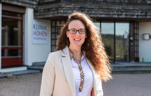 Frau vor Klinikgebäude der Klinik Hohenfreudenstadt, lächelnd in beiger Jacke.