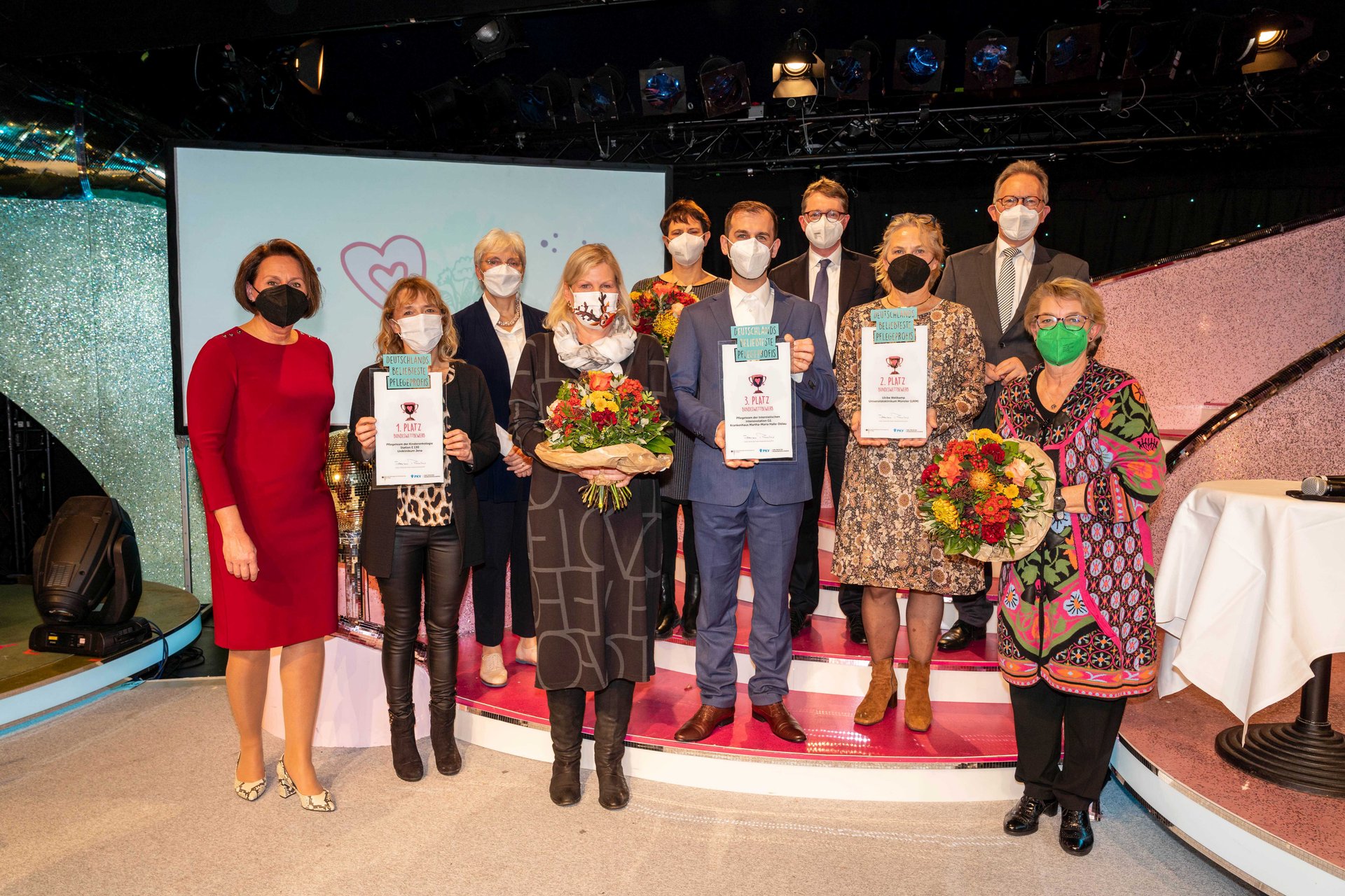 Menschen mit Masken halten Auszeichnungen bei einer Preisverleihung im Innenraum.