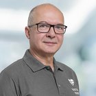 Porträt eines Mannes mit Brille und grauem Polohemd vor unscharfem Hintergrund.