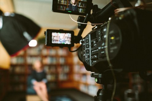 Kameraaufnahme eines Interviews vor Bücherregal, Fokus auf Kamera, unscharfe Person im Hintergrund.