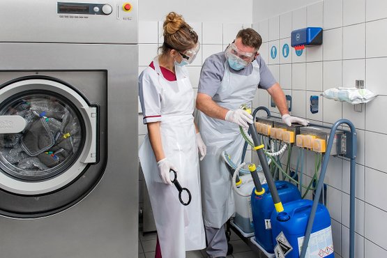 Zwei Personen mit Schutzkleidung beim Umgang mit Reinigungsmitteln in Wäscherei.