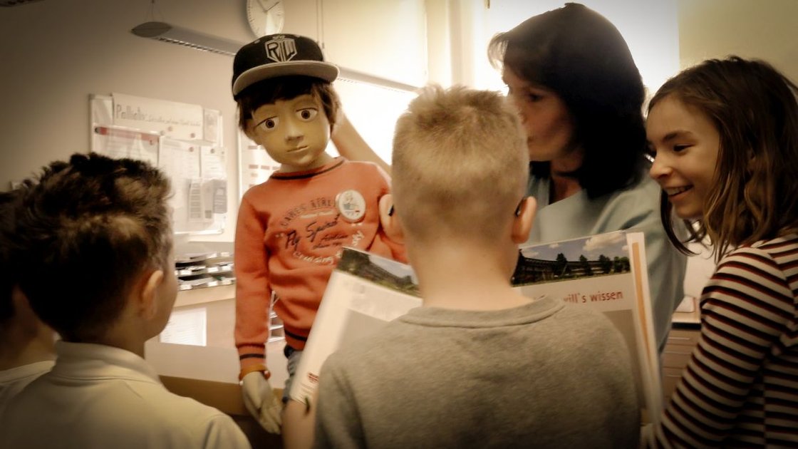 Kinder betrachten Puppe Jonas während eines Krankenhausprojekts, nahe einer Erwachsenen.