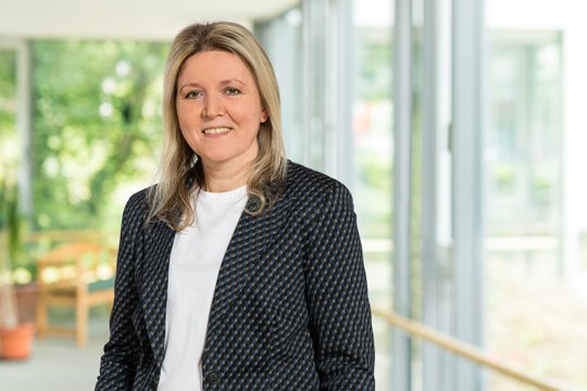 Renata Petrova lächelt in einem sonnigen Flur mit Pflanzen im Hintergrund.