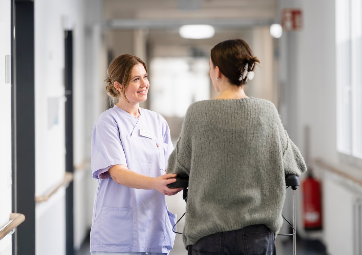 Pflegekraft unterstützt Patientin mit Gehhilfe im Krankenhausflur.