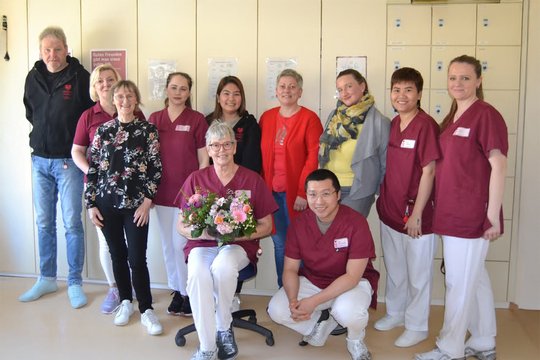 Neun Krankenhausmitarbeiter lächeln in die Kamera, eine Person hält Blumenstrauß.