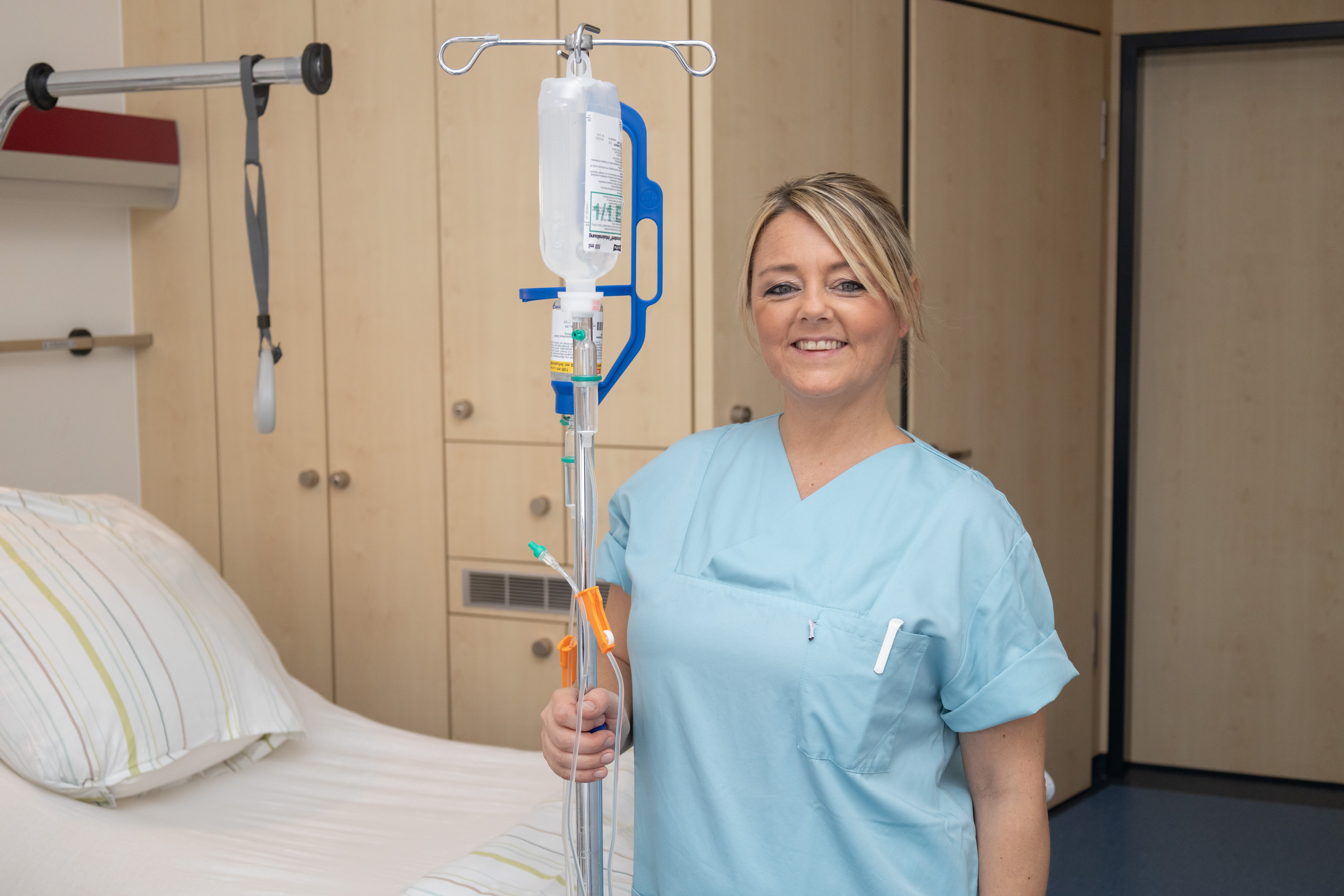 Pflegekraft in blauer Uniform mit Infusionsständer im Krankenhauszimmer.