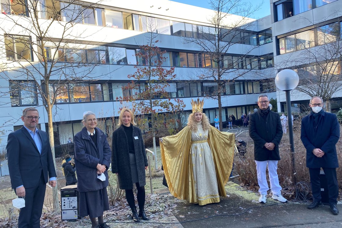 Gruppe von Menschen im Freien vor einem modernen Gebäude mit Christkind.