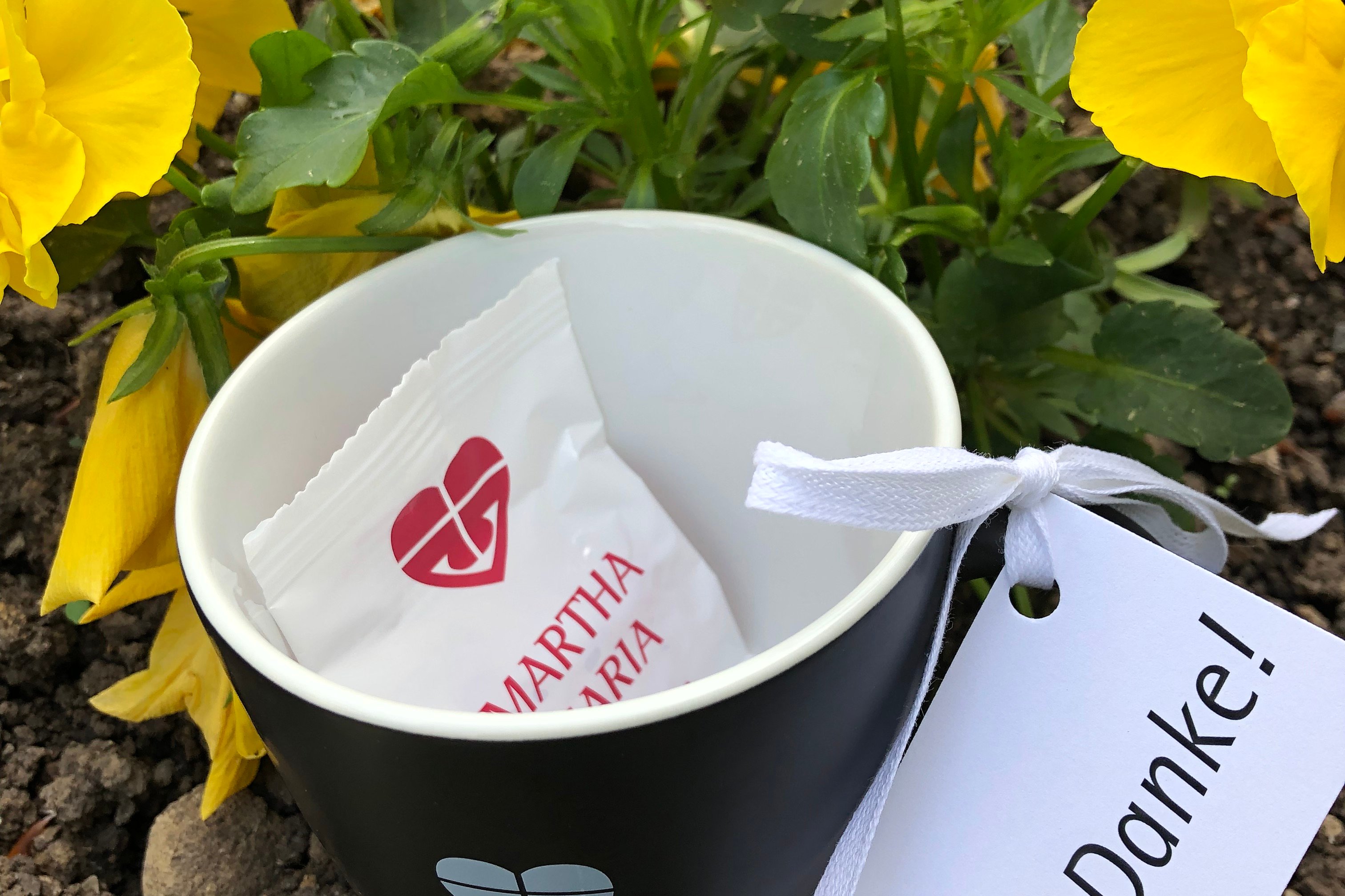 Schwarze Tasse mit 'Martha Maria'-Logo vor gelben Blumen, Dankeskarte angehängt.