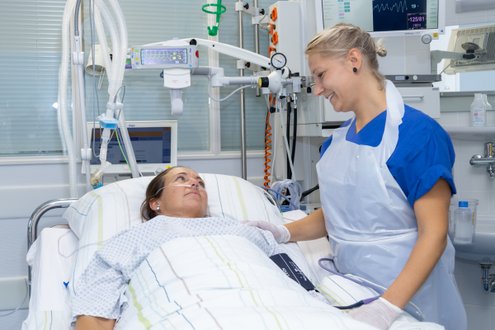 Krankenschwester lächelt Patientin im Krankenhausbett an, medizinische Geräte im Hintergrund.