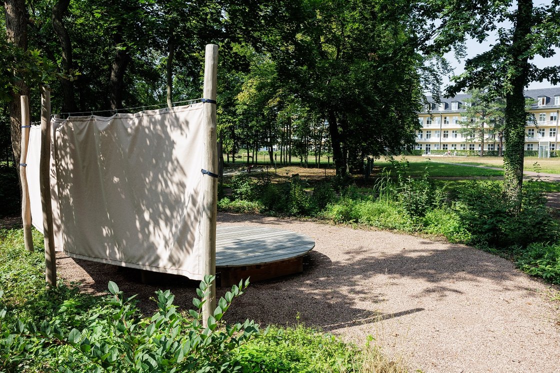 Holzplattform mit Stoffbahnen im besinnlichen Krankenhausgarten umgeben von Bäumen.