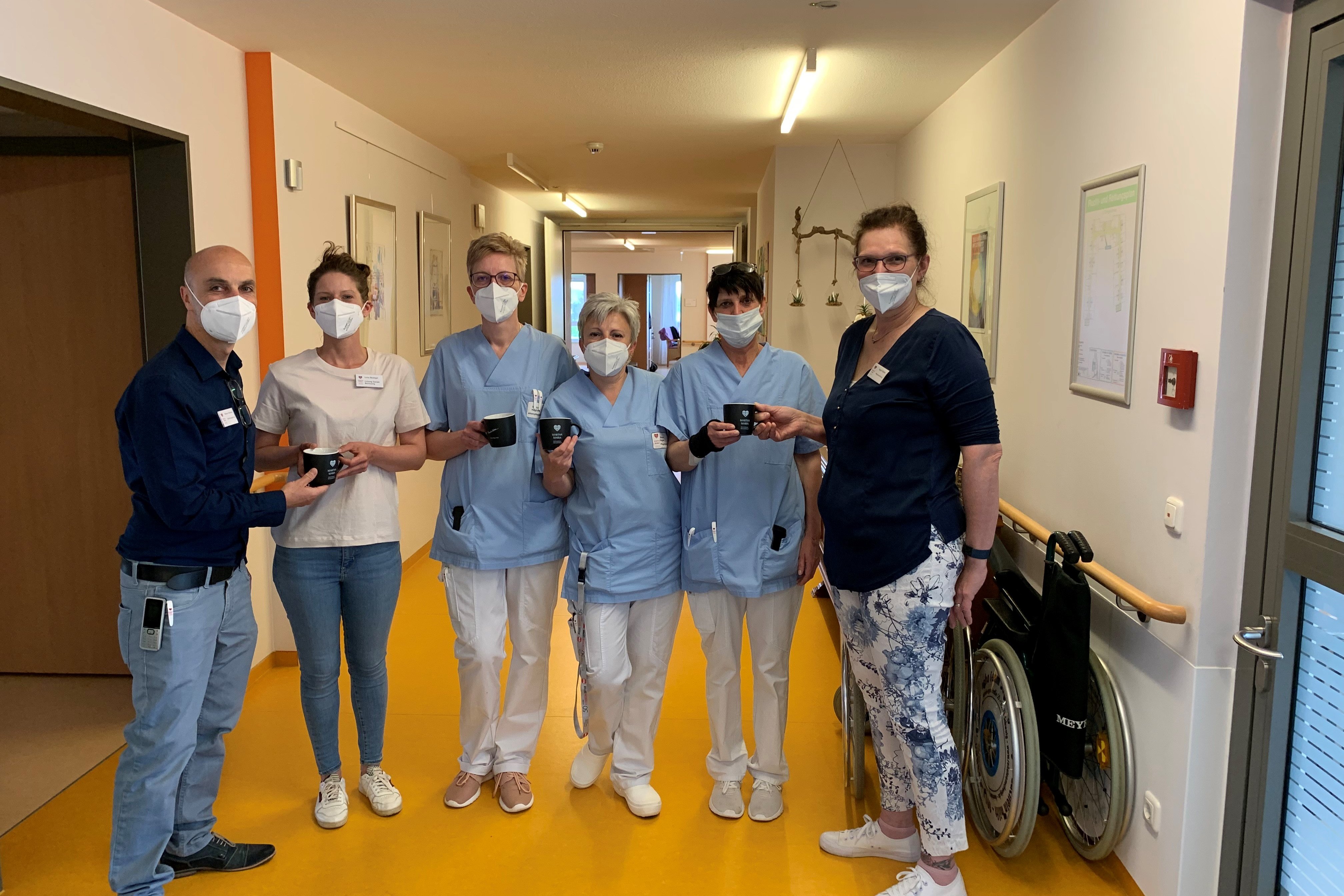 Pflegepersonal im Flur eines Pflegeheims mit Masken und Kaffeetassen.