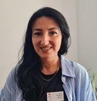 Frau mit Namensschild vor weißer Wand und Pflanze, lächelnd.