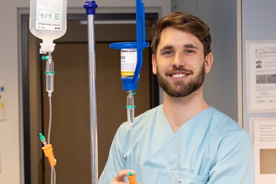 Männlicher Krankenpfleger in blauer Uniform lächelt, hält Infusionsständer im Krankenhaus.