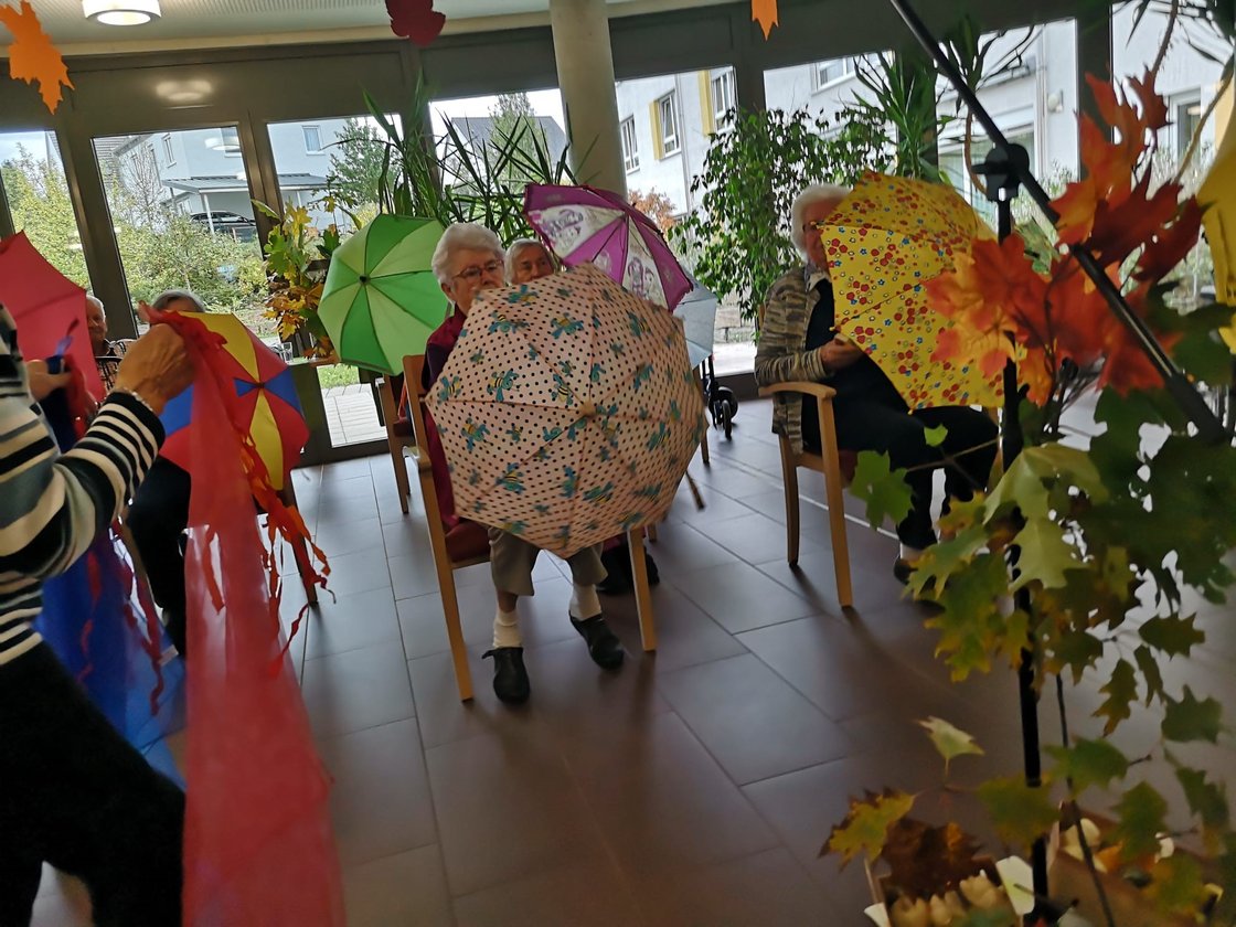 Senioren im Gemeinschaftsraum mit bunten Regenschirmen und Herbstdekoration.