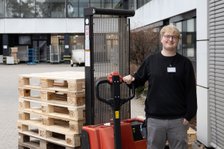 Mitarbeiter mit Hubwagen vor Palettenstapel in Lagerumgebung.