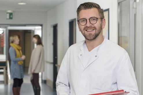 Lächelnder Arzt im Krankenhausflur mit zwei Personen im Hintergrund.