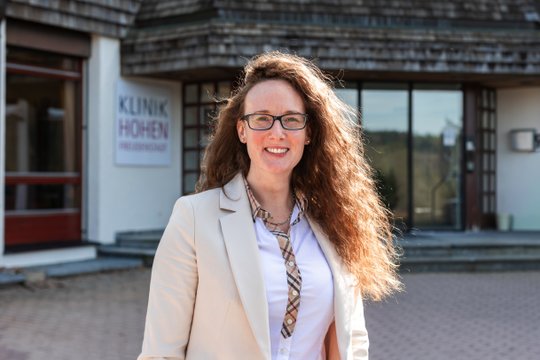 Frau vor Klinikgebäude der Klinik Hohenfreudenstadt, lächelnd in beiger Jacke.