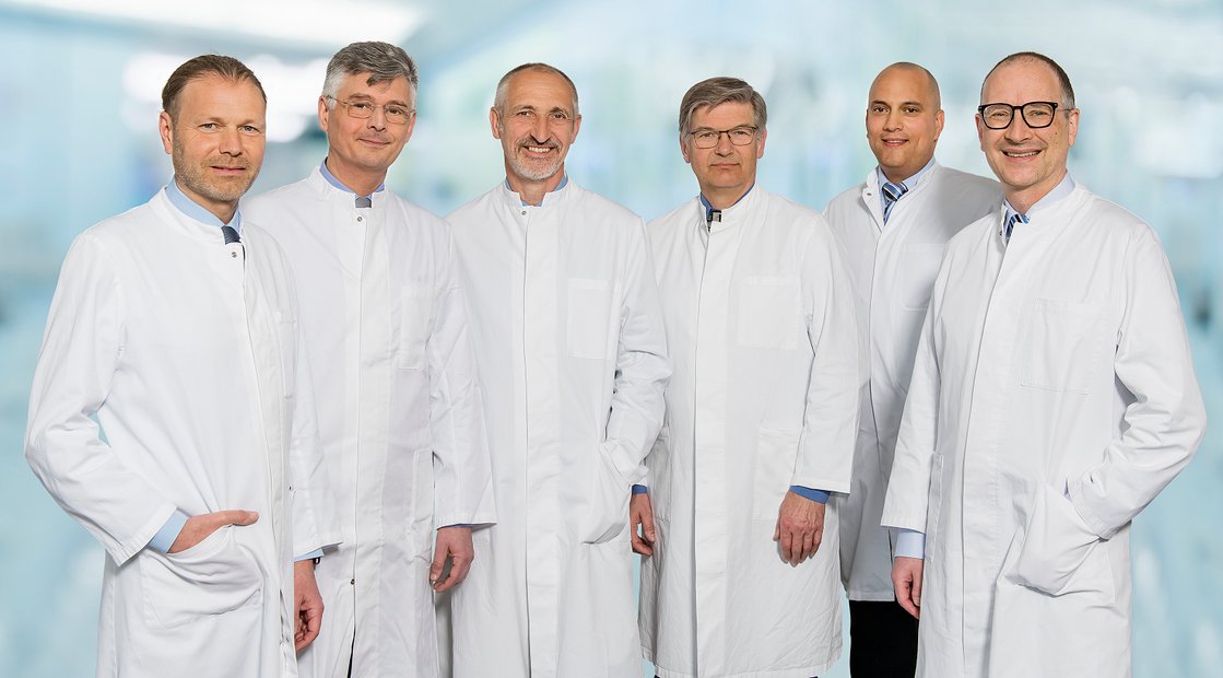 Sechs Ärzte in weißen Kitteln stehen in einer Krankenhausumgebung.