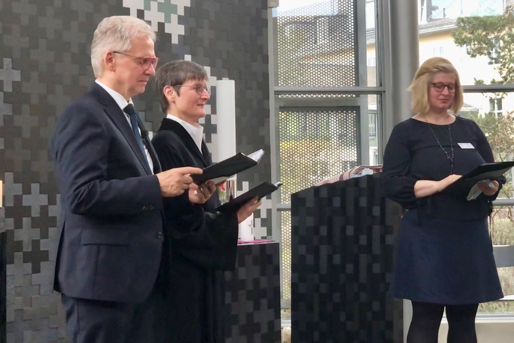Drei Personen bei einer formellen Veranstaltung mit Rednerpult und Notizbüchern.