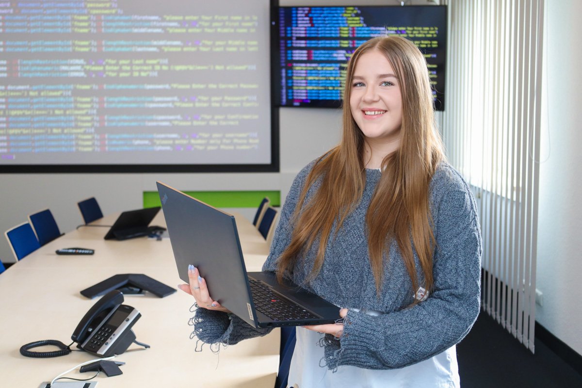 Junge Frau mit Laptop in Konferenzraum, zeigt Programmiercode auf Bildschirmen.