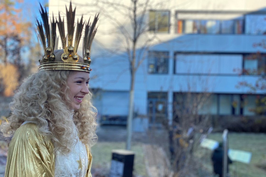 Frau in goldener Krone vor einem modernen Gebäude im Freien lächelnd.