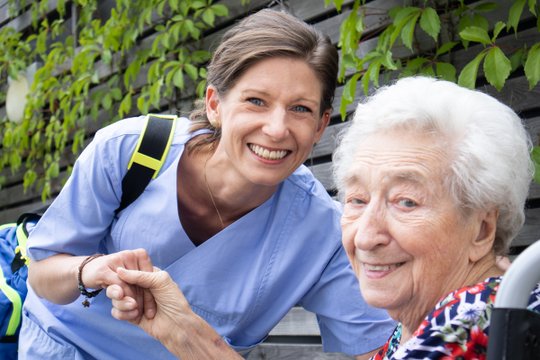 Pflegekraft in blauer Uniform lächelt mit älterer Dame im Garten.