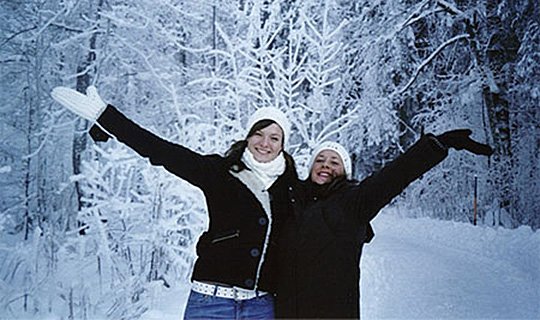 Zwei lächelnde Personen im Schnee, mit ausgestreckten Armen, umgeben von verschneiten Bäumen.