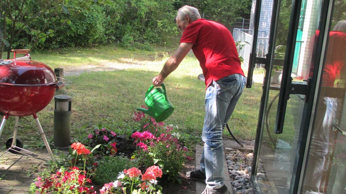 Älterer Mann im Garten gießt Blumen neben rotem Grill.