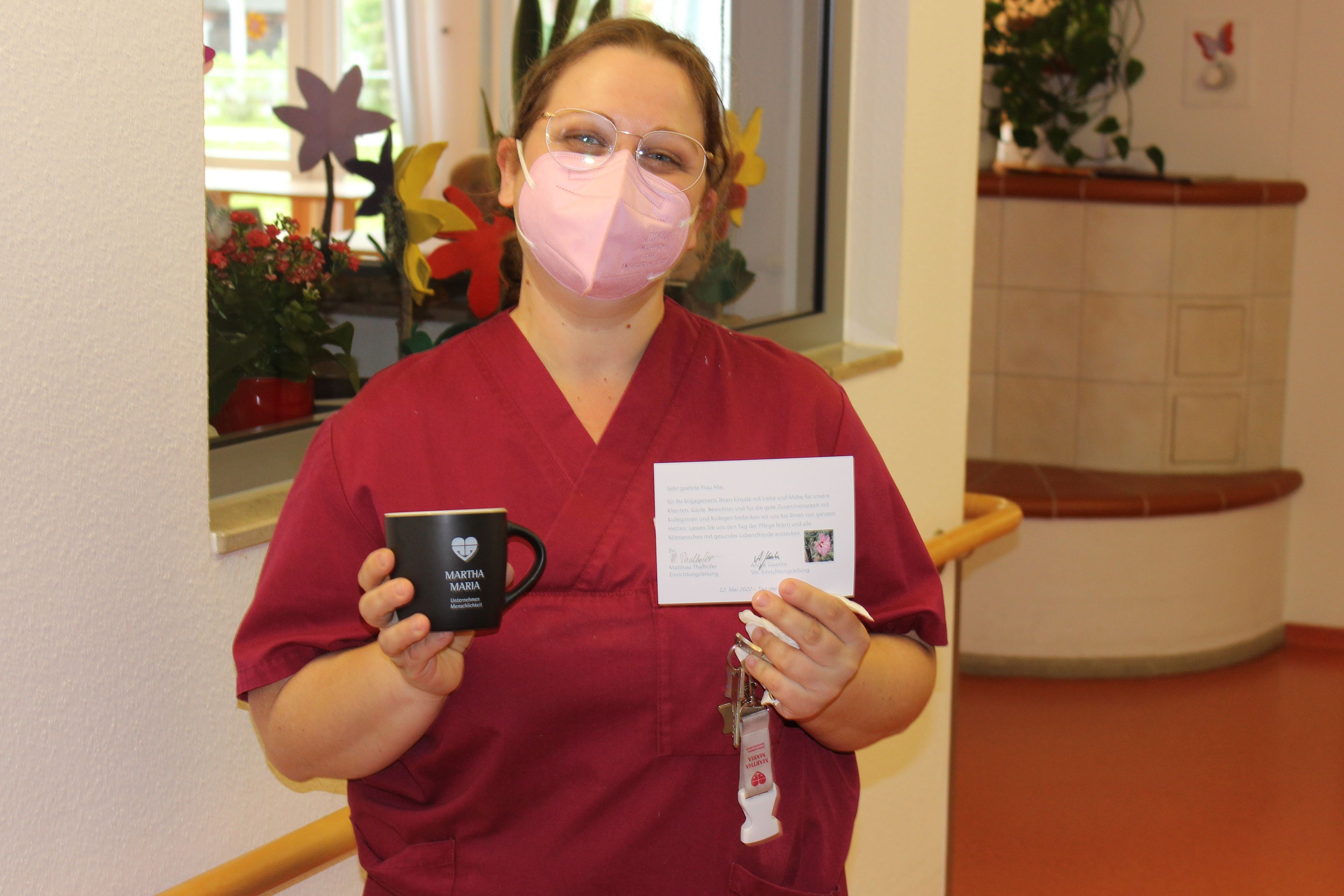 Pflegekraft in roter Uniform hält Tasse und Dankeskarte im Krankenhaus.
