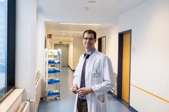 Arzt im weißen Kittel steht freundlich lächelnd im Krankenhausflur.