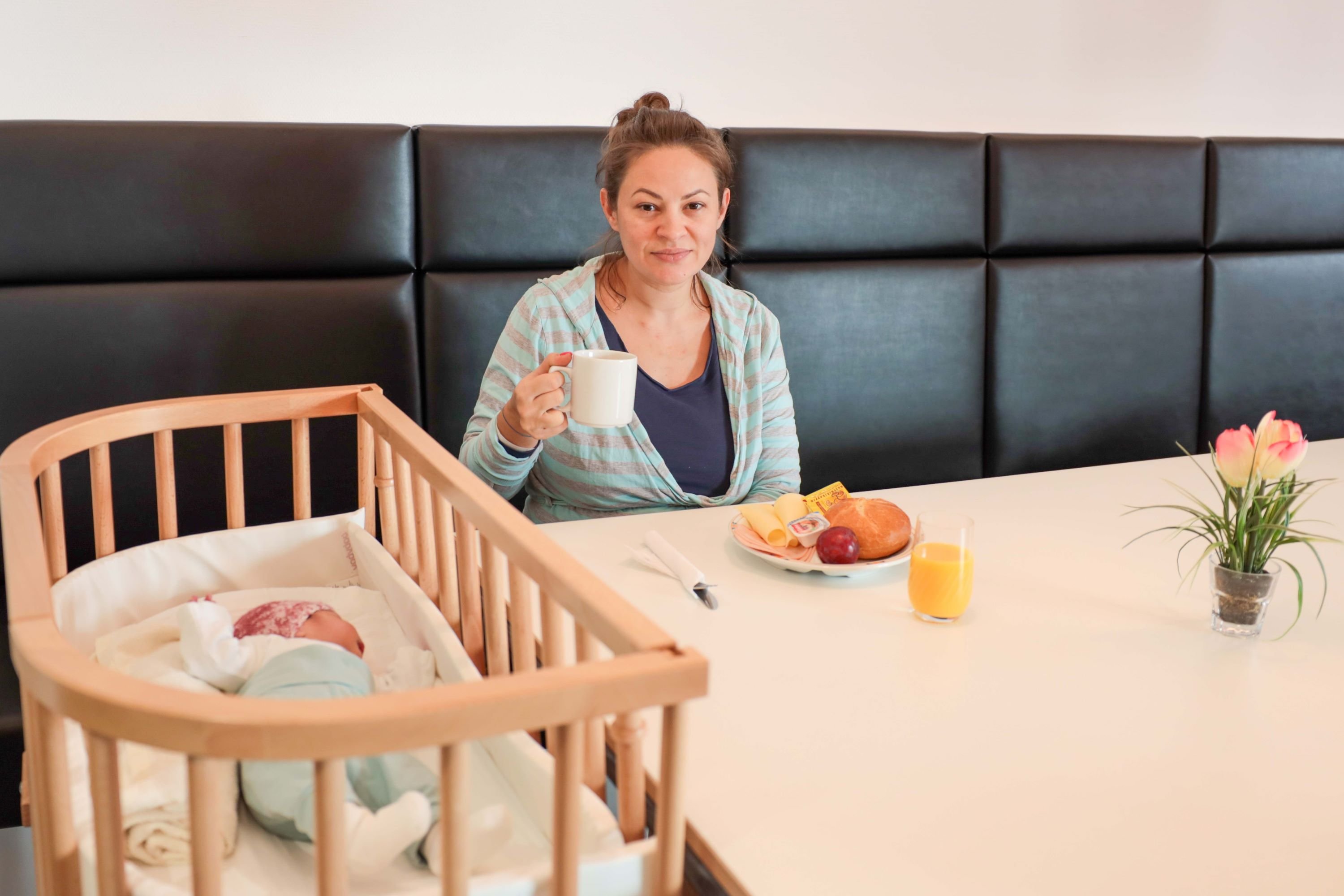 Mutter genießt Frühstück neben schlafendem Neugeborenen im Krankenhauszimmer.