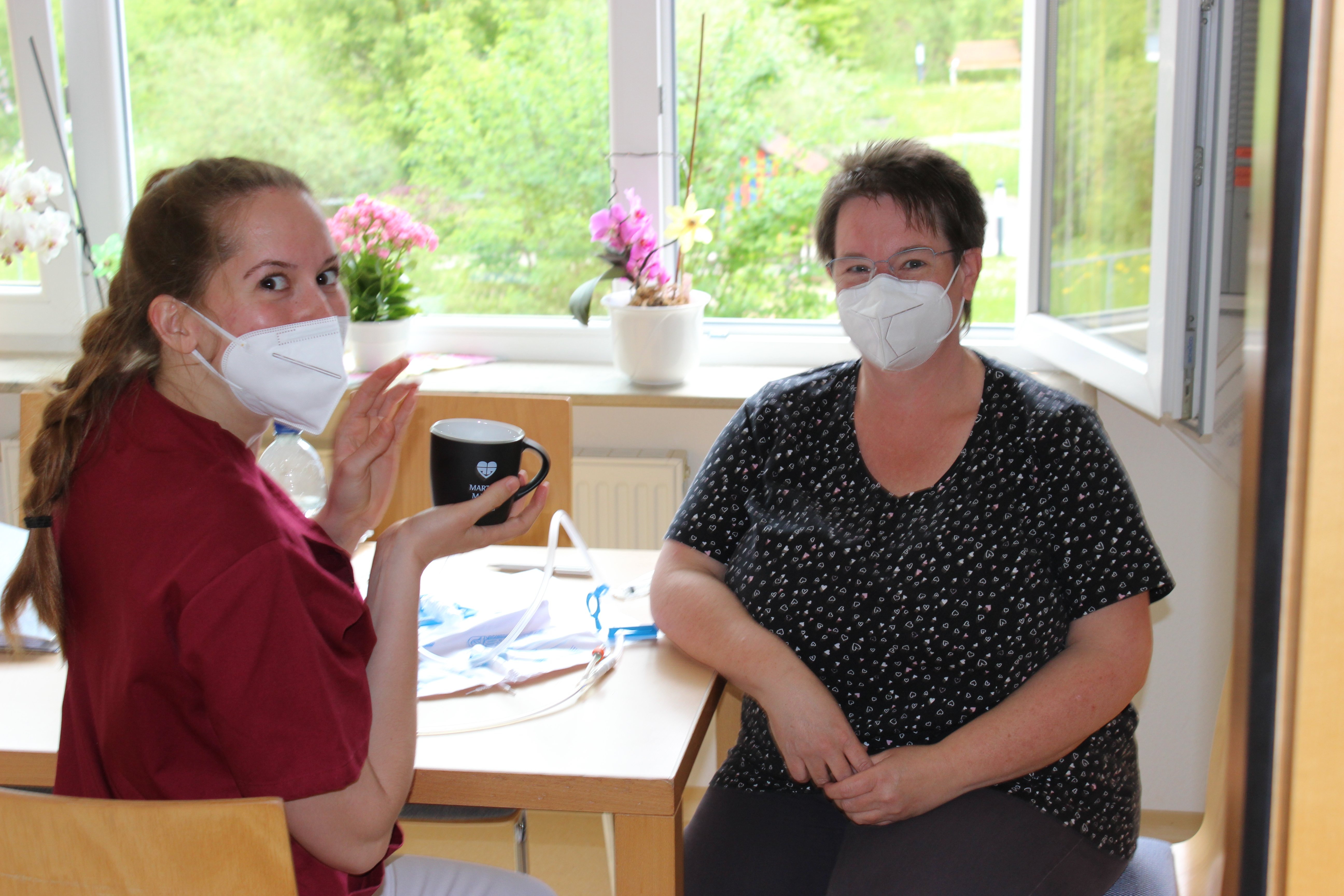 Zwei Frauen mit Masken im Gespräch an einem Tisch vor einem Fenster.