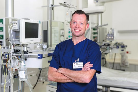 Arzt in blauer Kluft steht lächelnd in einem Krankenhauszimmer.