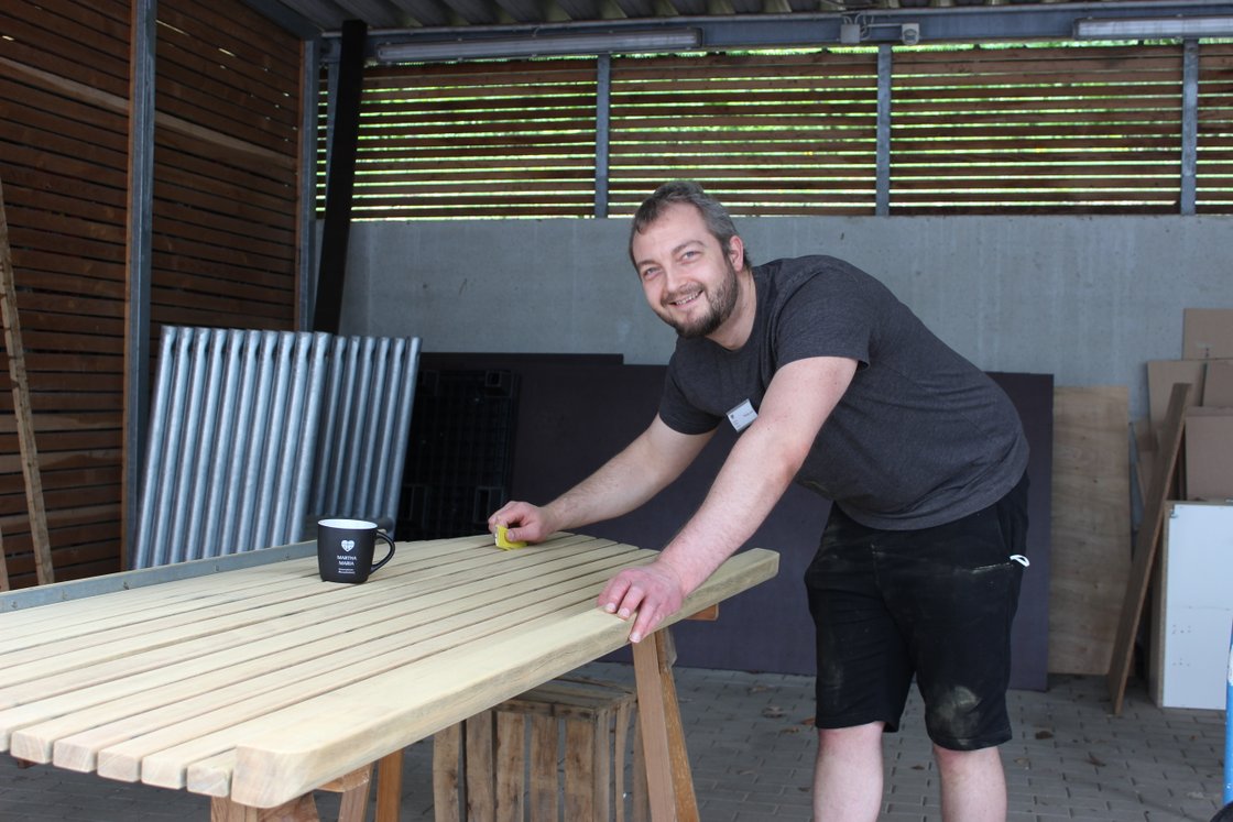 Mann schleift Holztisch in Werkstatt, lächelnd und mit Kaffeetasse.